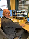 Mann sitter ved skrivebord med to skjermer, laptop og kontorutstyr ved et vindu med persienner.