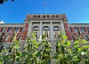 Røde murbygninger med Høyesterett i midten, sett bak høye grønne planter mot blå himmel.