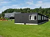 Et moderne, mørkt skolebygg står på en grønn plen med skogkledd bakgrunn og blå himmel.