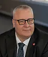 Portrett av en mann med briller i mørk dress og slips foran en nøytral bakgrunn.