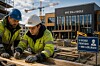 To arbeidere i verneutstyr arbeider med treverk på en byggeplass foran en skole som bygges.