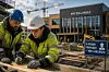 To arbeidere i verneutstyr arbeider med treverk på en byggeplass foran en skole som bygges.