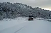 Snøplog kjører på en vinterdekket vei foran snødekte trær og åser.