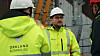 To arbeidere med hjelm og refleksjakker står foran stillas og bygningsmaterialer på en byggeplass.