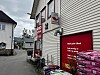 Dagligvarebutikk med røde skilt og varer utstilt ved en smal gate mellom bygninger.
