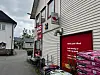 Dagligvarebutikk med røde skilt og varer utstilt ved en smal gate mellom bygninger.