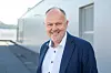 Smilende mann i blå blazer står foran en moderne bygning utendørs.