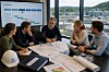 Fem personer sitter rundt et bord med tegninger, dokumenter og hjelm i et møterom med store vinduer og utsikt til et moderne VA-anlegg.