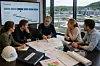 Fem personer sitter rundt et bord med tegninger, dokumenter og hjelm i et møterom med store vinduer og utsikt til et moderne VA-anlegg.