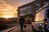 En person går med verktøykasse mot et moderne kontorbygg ved solnedgang, med en varebil parkert til høyre.