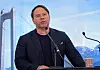 Mann taler ved en talerstol foran en bro og snødekte fjell.