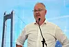 Mann i hvit skjorte taler i mikrofon foran et bilde av en hengebro.
