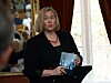 Kvinne ved talerstol holder en blå publikasjon i et rom med vindu og gardiner.