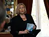 Kvinne ved talerstol holder en blå publikasjon i et rom med vindu og gardiner.