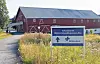 Blå Kors-skilt ved innkjørselen til et rødt gårdsanlegg med vegetasjon rundt.