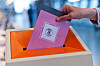 Hånd legger en rosa stemmeseddel i en oransje valgurne.