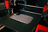 Nærbilde av en 3D-printer som lager et grått, lagdelt objekt på en svart byggeplate.