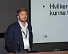 Mann ved podium foran en stor presentasjonsskjerm med tekst.