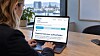 Person ved laptop på kontor med nettside på skjermen og byutsikt i bakgrunnen.