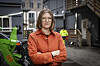 Kvinne i oransje arbeidsjakke foran anleggsmaskiner og arbeidere i synlighetsklær.