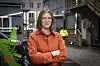 Kvinne i oransje arbeidsjakke foran anleggsmaskiner og arbeidere i synlighetsklær.