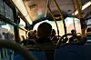 Passasjerer sett bakfra i en solfylt lokalbuss med bylandskap utenfor vinduene.