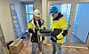 To arbeidere i hjelmer og verneklær arbeider sammen i et rom under oppussing.