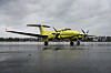 Gult tomotors turbopropfly på våt asfalt under grå skyer, med skog i bakgrunnen.