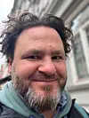 Smilende skjegget mann i hettegenser fotografert på en bygategate.