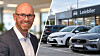 Mann i dress ved siden av flere biler på en bilutleieplass med skiltet «Leiebiler».