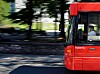 En rød Ruter-buss kjører langs en vei med trær og steinmur i bakgrunnen.